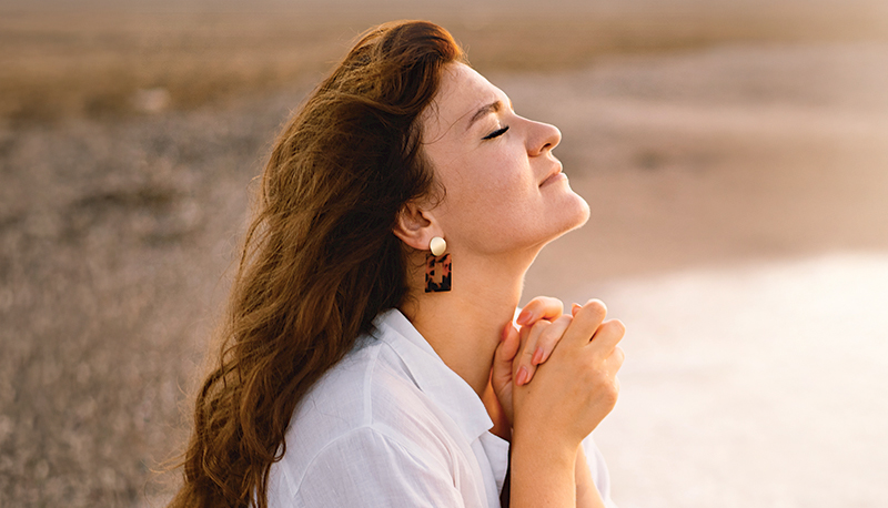 Woman closed her eyes, praying on a sea during beautiful sunset. Hands folded in prayer concept for faith, spirituality and religion.