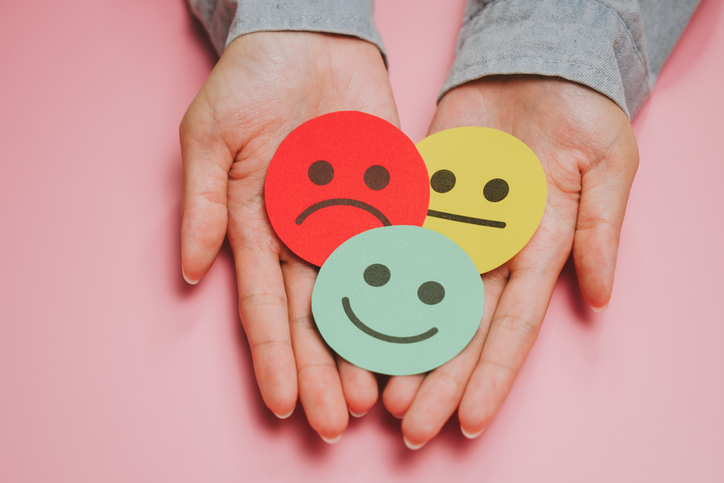 Colorful emotion faces on paper held in hands, representing happy, neutral, and sad feelings with soft pink background creating thoughtful and expressive mood