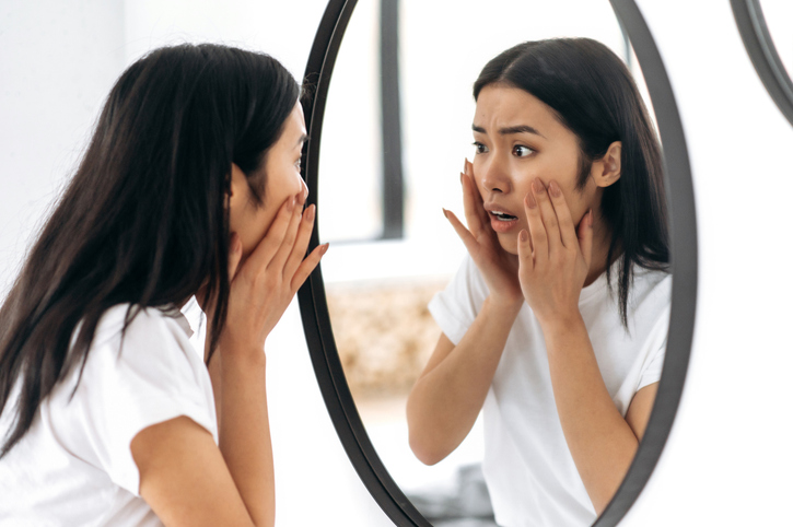 Stunned worried Asian brunette girl, stands at home in front of a mirror, examines the skin of her face, unhappy with the condition of the skin, needs cosmetology services, moisturizing, care
