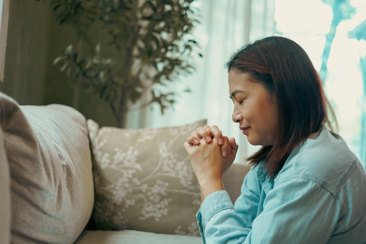 ​Woman sits on a knee and prays in front of a sofa.