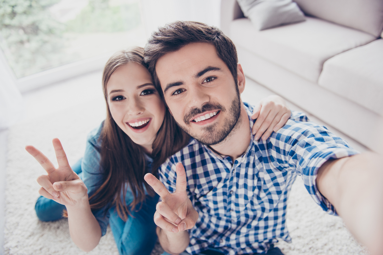 Cheerful playful sister and brother are taking selfie and showing peace signs. They are at home indoors, in casual outfits. They both have beaming smiles